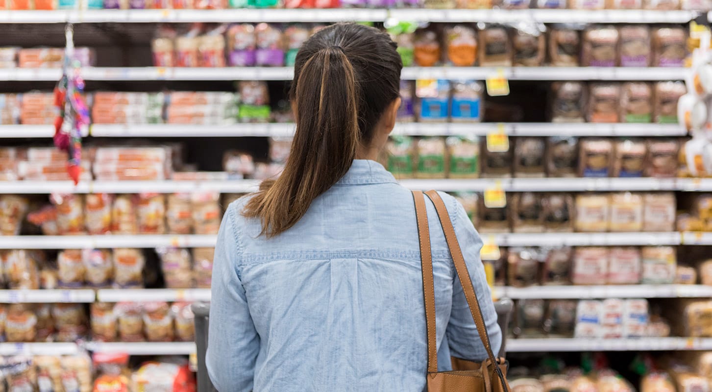 A woman with a ponytail, wearing a denim shirt and brown shoulder bag, stands in front of shelves of bread in a grocery store.